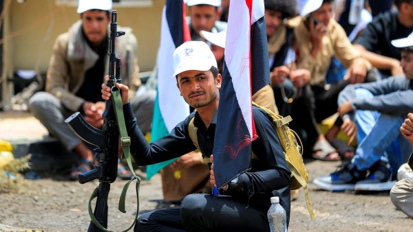 Un homme armé, tenant un drapeau, assis parmi un groupe de personnes en arrière-plan.