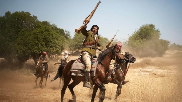 Des cavaliers armés avancent sur un terrain poussiéreux, dans un décor rural.