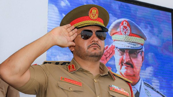 Un homme en uniforme militaire, lunettes noires, salue devant un écran.
