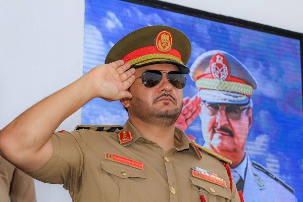 Un homme en uniforme militaire, lunettes noires, salue devant un écran.