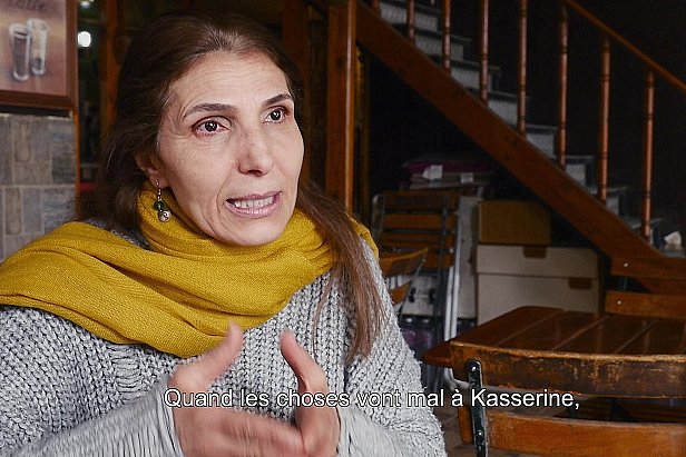 L'image montre une femme en train de parler, assise dans un environnement intérieur. Elle porte un vêtement gris avec un foulard jaune autour du cou. L'arrière-plan présente des escaliers en bois et des tables en bois, ce qui donne une ambiance chaleureuse et intime. Elle semble s'exprimer avec passion, évoquant probablement des enjeux importants liés à la ville de Kasserine.