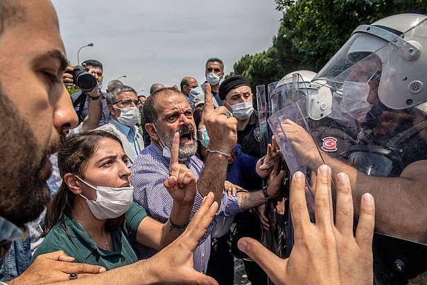 تظهر الصورة محتجين يتجمعون في مكان عام، حيث يبدو أن هناك توترًا في الأجواء. بعض الأشخاص يحملون لافتات، بينما يتواجد عدد من رجال الشرطة الذين يرتدون خوذات ودروع لمواجهة الحشد. يبدو أن هناك تفاعلًا حادًا بين المتظاهرين ورجال الأمن، مع تعبيرات وجهية تشير إلى الغضب والاحتجاج. بعض الأشخاص يرتدون أقنعة للوجه، مما يدل على احتمال وجود تدابير صحية. الأشجار والخضرة، تشير إلى أن الموقع في منطقة حضرية.